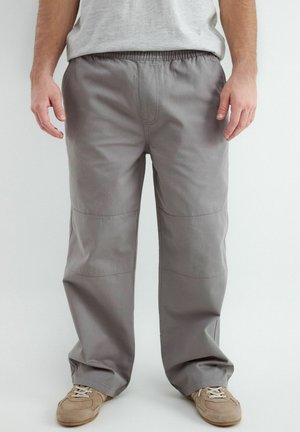 Mężczyzna noszący luźne szare spodnie i beżowe trampki, stojący na jasnym tle.