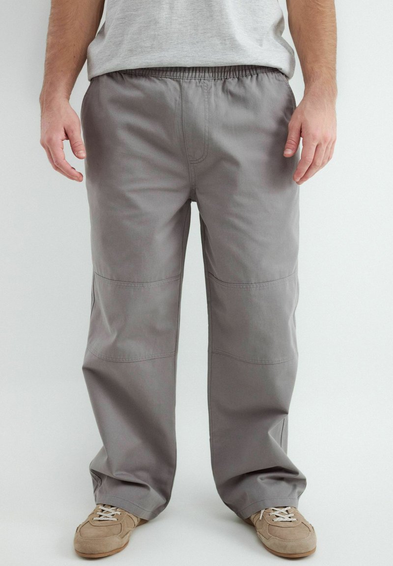 Homme portant un pantalon gris ample et des baskets beiges, debout sur un fond clair.