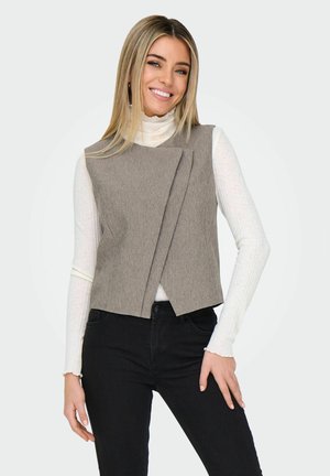 Žena s dlouhými blond vlasy má na sobě šedou asymetrickou bezruko vestu přes bílý žebrovaný rolák a černé kalhoty, usmívá se a má ruku na boku.