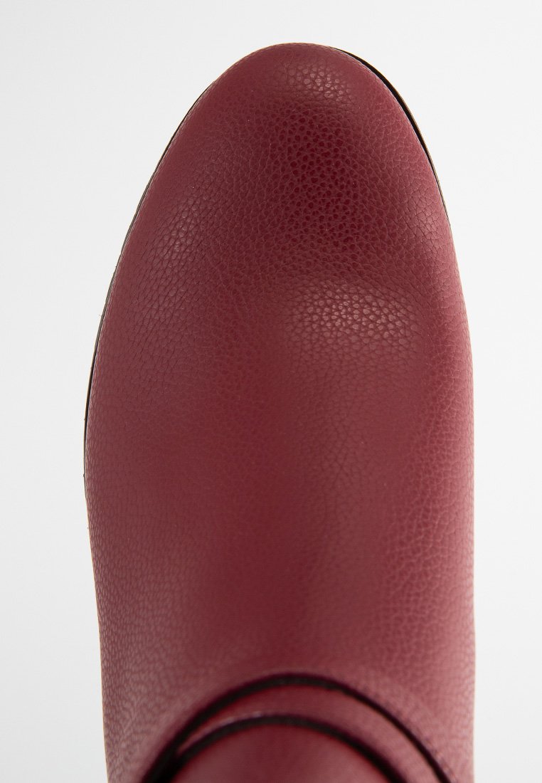 Rote Lederstiefelette mit einer genarbten Oberfläche, runder Zehenpartie und kreisförmigem Akzentdetail am Knöchel. Glatte Sohle mit minimaler Naht.