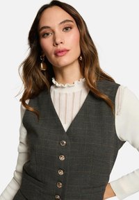 Gilet a quadri scuro con cinque bottoni sul davanti indossato su un dolcevita bianca a coste. Presenta un collo delicatamente arricciato e una texture morbida.