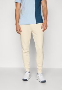 Light beige sweatpants with a slim fit, made from soft material. Paired with a color-blocked blue and navy short-sleeve shirt and white sneakers.