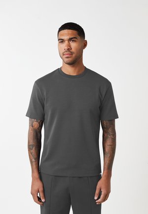 Homme aux cheveux courts et tatouages portant un t-shirt à manches courtes gris foncé et un pantalon assorti sur fond blanc uni.