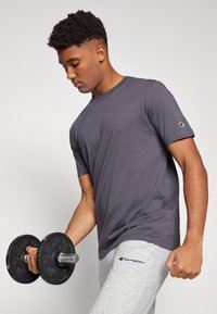 T-shirt gris à manches courtes en matériau doux, porté avec un pantalon de survêtement gris comportant un logo. La personne tient un haltère.
