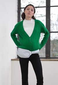 Cardigan verde de malha em ponto trançado com decote em V e fecho de botão, combinado com um suéter de gola alta branco e calças pretas, destacando uma mala de cintura branca.