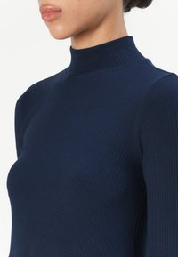Navy ribbed turtleneck top with long sleeves. Textured fabric with a fitted shape and standing collar, highlighting a sleek design.