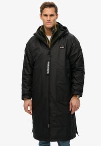 Zwarte lange jas met een rits aan de voorkant, grote zakken en een verstelbare capuchon. Beschikt over een gestructureerde stof en een logo op de borst.