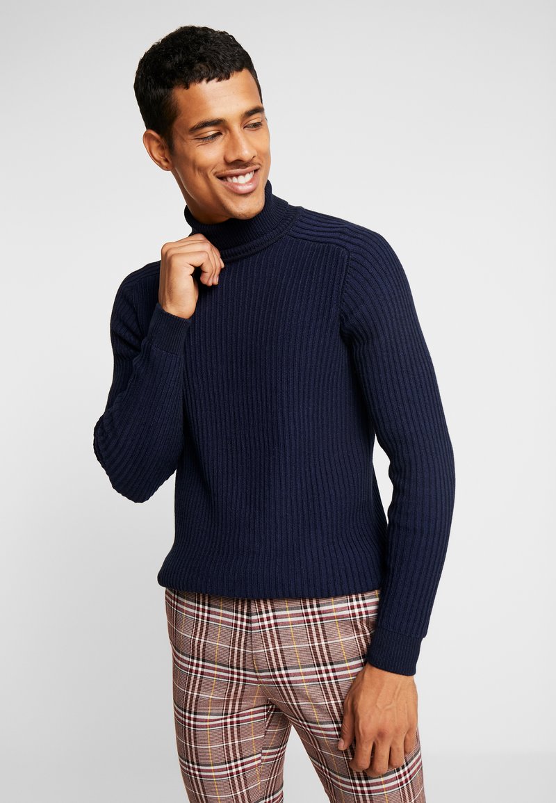 Pull à col roulé côtelé marine avec des manches longues et des poignets ajustés, associé à un pantalon à carreaux avec des accents marron, crème et rouge.