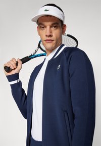 Casaco azul-marinho com fecho éclair e detalhes brancos, em tecido leve, usado por cima de uma polo branca, segurado por uma pessoa que exibe uma raquete de ténis.