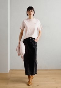 T-shirt rosa chiaro, gonna di jeans nera, stivali beige alla caviglia. La t-shirt presenta un piccolo logo; la gonna è lunga con tasche frontali.