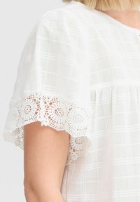 Blusa bianca con un motivo a griglia testurizzato, con maniche corte decorate da un intricato pizzo lungo i bordi.