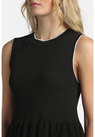 Femme portant une robe noire côtelée sans manches avec un liseré blanc autour des emmanchures et du col, montrant les épaules et une partie du cou.