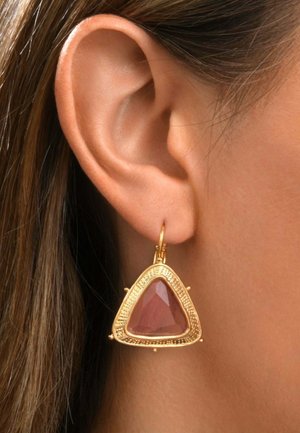 Gros plan sur l'oreille d'une personne portant une boucle d'oreille triangulaire en or avec une pierre rouge-brun facettée au centre.