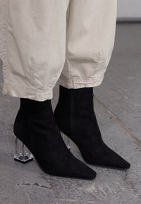 Botas de tobillo de terciopelo negro con puntas afiladas y tacones geométricos transparentes. Las botas cuentan con un cierre de cremallera lateral.