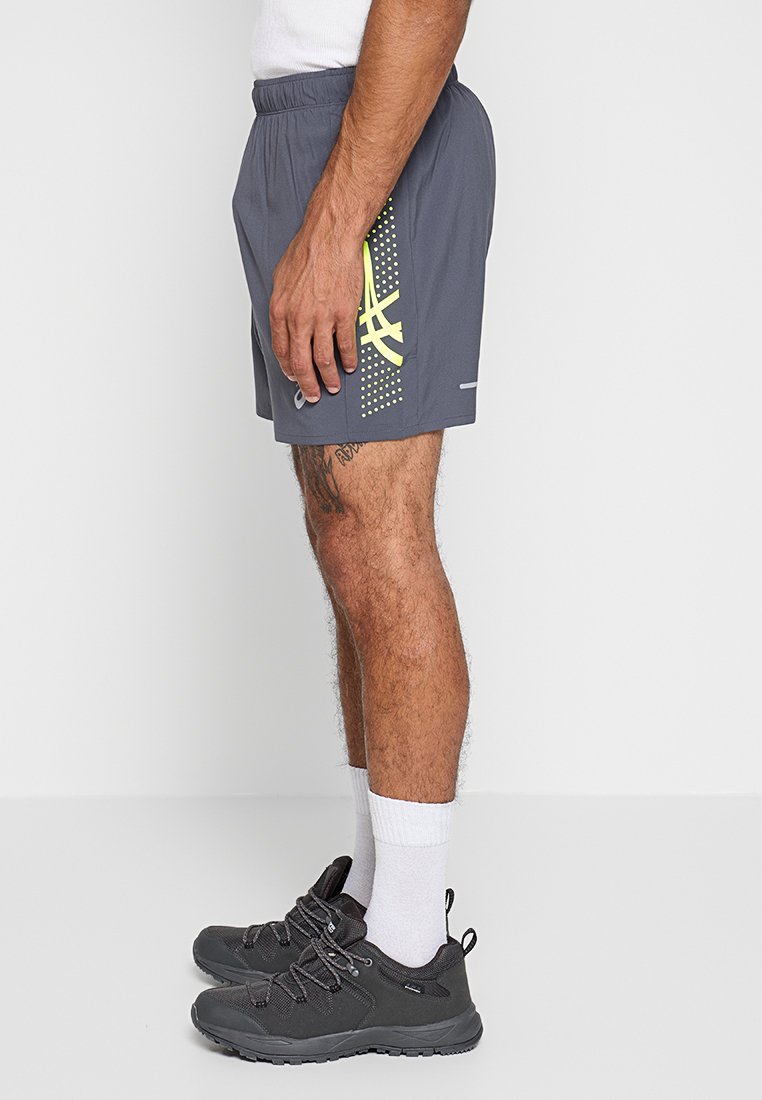 Man in grijze sportshorts met een geel logo, witte sokken en zwarte hardloopschoenen, staand in profiel tegen een witte achtergrond.