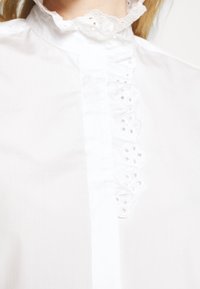 Weißes Hemd mit hohem Rüschensaumkragen und dekorativer Spitzenstickerei, mittigem Knopfverschluss und glatter Textur.