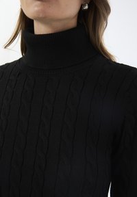 Nahaufnahme einer Person, die einen schwarzen, grob gestrickten Rollkragenpullover trägt, mit langen braunen Haaren vor einem schlichten weißen Hintergrund.