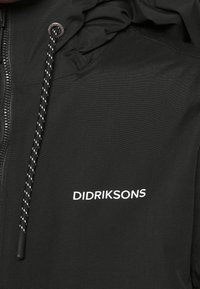 Zwarte jas met capuchon van gestructureerde stof. Voorzien van een ritssluiting met een zilverkleurige trekker en een wit logo op de borst gedrukt. Trekkoord met zwart-wit patroon.