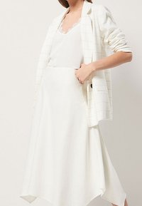 Blazer bianco con motivo a griglia e maniche arrotolate, indossato sopra una canotta bianca, abbinato a una gonna bianca fluida con orlo asimmetrico.