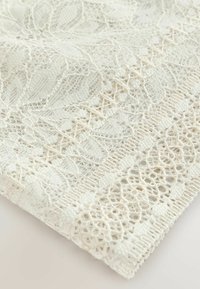 Un tissu en dentelle blanche détaillée présentant des motifs floraux et géométriques avec un bord festonné sur un fond clair.