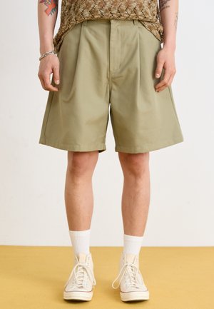 Shorts kaki plissés avec poches latérales, fabriqués en tissu lisse. Associés à des baskets montantes blanches et des chaussettes côtelées.