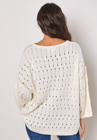 Leichter, oversized, cremefarbener Pullover mit offenem Strickmuster, großen Ärmeln und weit ausgeschnittenem Hals. Mit bunten Flecken durchzogen.