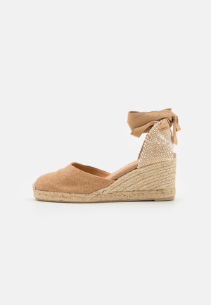 Espadrille compensée avec tige en tissu beige, semelle en jute tressée, lien à nouer autour de la cheville et contrefort tissé dans des tons neutres sur fond blanc.