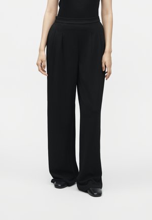 Vrouw draagt een zwarte high-waisted broek met wijde pijpen en zwarte schoenen, staand tegen een effen witte achtergrond.