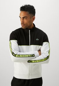 Juoda, balta ir neoninė žalia zip-up sportinė striukė. Ypatybės: aukštas kaklas, kontrastingos juostelės ir logotipo detalė ant krūtinės bei rankovių.