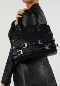 Czarna skórzana torebka o zróżnicowanej strukturze, z złotymi akcentami, ćwiekowanymi paskami i podwójnymi uchwytami. Na przodzie znajduje się emblem.