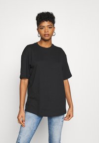 T-shirt nera in cotone con maniche corte, colletto a coste e un sottile logo in rilievo sul davanti. Abbinata a jeans strappati blu chiaro.