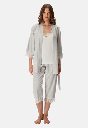 Mujer de pie descalza con bata gris claro con encaje y pantalones sobre un camisón blanco con encaje, sobre un fondo sencillo.