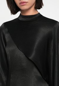 Φόρεμα από μαύρο σατέν ύφασμα με ψηλό γιακά και ασύμμετρο σχέδιο, με λεία υφή και διακριτική λάμψη.