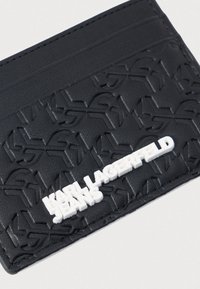 Porta-cartões de couro preto com padrão de logotipo em relevo. Apresenta um destaque de logotipo branco e três slots para cartões na parte superior. Superfície texturizada.