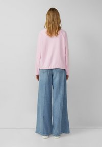 Jasnoróżowa bluzka z długim rękawem o zaokrąglonym dole, zestawiona z szerokimi jasnoniebieskimi dżinsami. Oba elementy garderoby mają gładką teksturę.