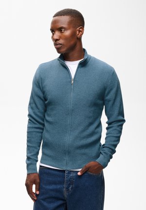 Homme portant un pull zippé bleu sur un t-shirt blanc et un jean bleu, debout avec une main dans la poche, regardant sur le côté.