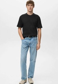 Mlad moški stoji oblečen v črno majico, svetlo modre kavbojke s črnim pasom, bež superge, na enobarvni beli ozadju.