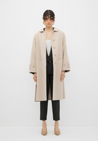Cappotto di lana beige con ampio rever e sei bottoni decorativi, abbinato a una maglia bianca e pantaloni a righe sottili. Texture liscia, design sartoriale.