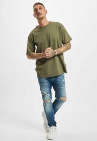 Tatuiruotes turintis vyras, vilkintis alyvuogių žalumo spalvos marškinėlius, suplyšusius mėlynus džinsus, baltus sportinius batus, stovi su sukryžiuotomis rankomis ir viena koja perrišta.