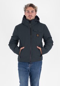 Ciemna, ocieplana kurtka z kapturem, z zamkiem błyskawicznym i kieszenią na piersi. Materiał jest gładki, z brązowymi akcentami przy kieszeniach.