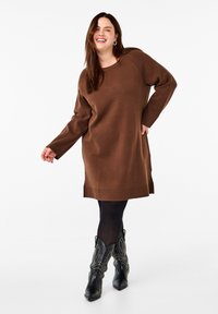 Ruskea neulemekko, jossa on pitkät hihat, rento malli, ribbaus hihansuissa ja helmassa, yhdistettynä teksturoituihin mustiin cowboy-kengiin, joissa on niittejä.
