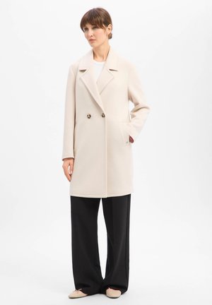 Frau mit beigem doppeltreihigem Mantel mit Schildpattknöpfen, schwarzen weit geschnittenen Hosen und beigen flachen Schuhen, die vor einem weißen Hintergrund steht.