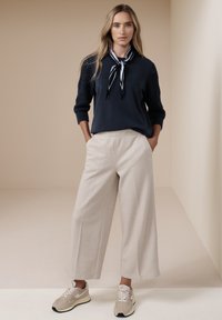 Donna in piedi con le mani nelle tasche, indossa un maglione blu navy, una sciarpa a righe intorno al collo, pantaloni beige a gamba larga e sneakers beige su sfondo neutro.