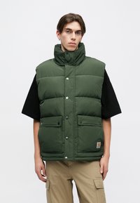 Pehmea roheline vest, millel on kõrge krae, nööbid ja kaks eesmist taskut. Kantud musta T-särgi kohal, koos beežide cargo-pükstega.