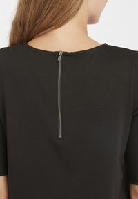 Maglietta nera con scollatura rotonda e maniche corte. Presenta una cerniera argentata visibile sul retro, che aggiunge un tocco di dettaglio al design semplice.