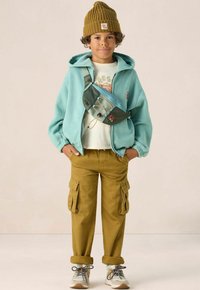 Sweatshirt turquoise, t-shirt à motif beige, sac banane vert olive, pantalons cargo moutarde et baskets grises avec accents marron.