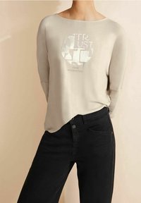 Helles beiges Langarmoberteil mit rundem Halsausschnitt und Grafikdruck, kombiniert mit schwarzen Jeans im klassischen Schnitt und mit dezenten Nähten.