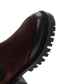 Gros plan d'une botte en daim marron avec bout en cuir noir et semelle épaisse à crampons sur fond blanc.