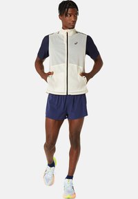 Letvægts, ærmeløs cremevest med marineblå ærmer, forlommer og høj krave. Matchet med marineblå shorts og multifarvede løbesko.