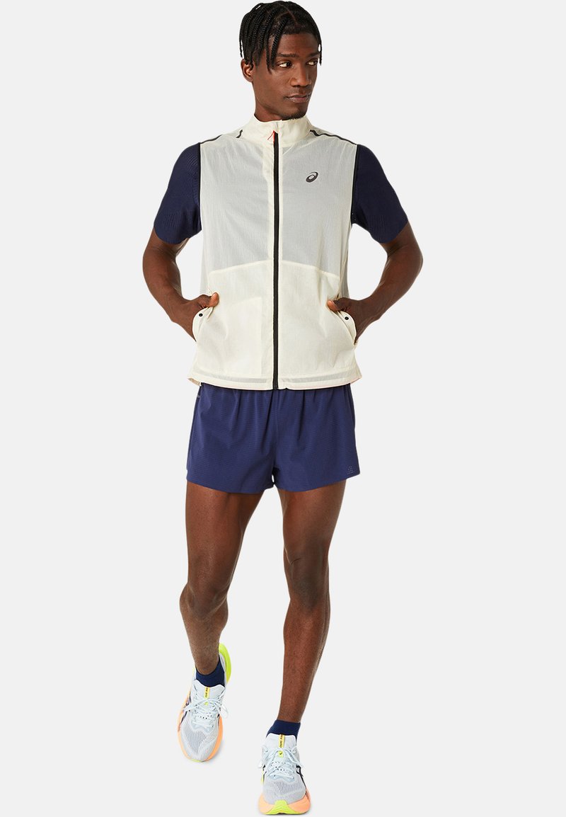 Letvægts, ærmeløs cremevest med marineblå ærmer, forlommer og høj krave. Matchet med marineblå shorts og multifarvede løbesko.